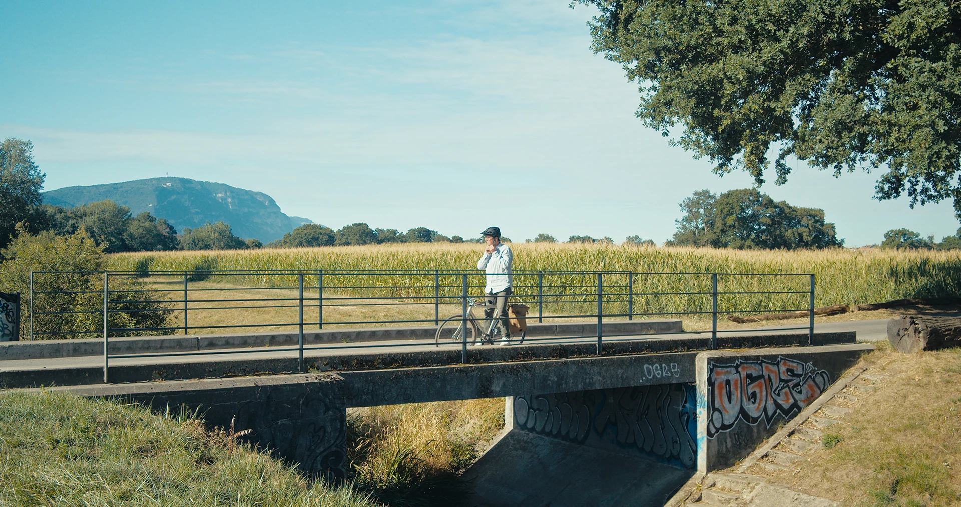 Cornland Studio - Un cycliste arrêté sur un pont dans la campagne genevoise enlève son casque.